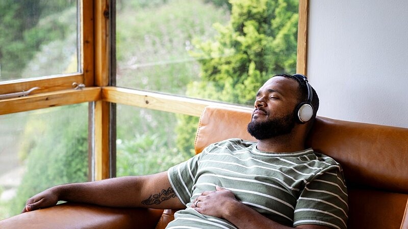 Eine Person liegt entspannt auf einem Sofa, hört mit Kopfhörern Musik und blickt in Richtung Fenster – symbolisch für Selbstfürsorge, Ruhe und bewusste Erholung., Bild: SolStock / gettyimages